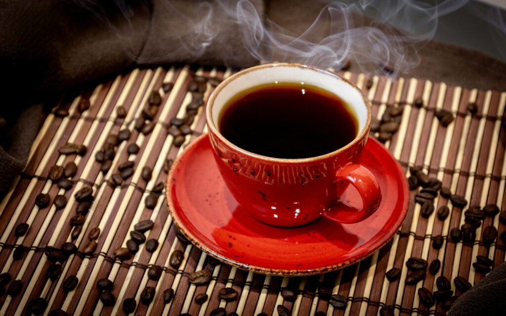 A steaming cup of coffee in a red mug surrounded by coffee beans on a bamboo mat.