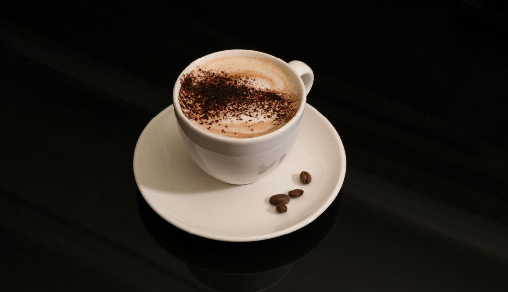 Cup of cappuccino topped with cocoa powder on a saucer with coffee beans.