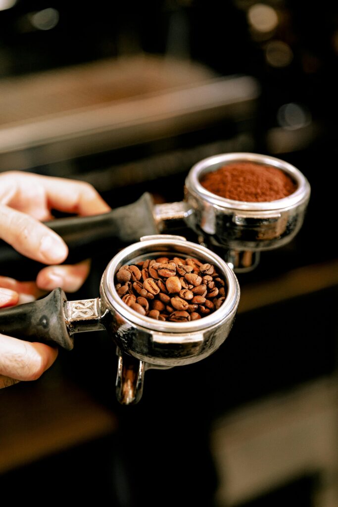 Close-up of a barista's hands holding portafilters filled with coffee beans and ground coffee.